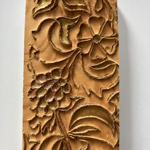 Pode incluir: Placa retangular de madeira com um desenho floral esculpido. O desenho apresenta uma flor, folhas e um cacho de uvas, com detalhes dourados. Uma pequena argola de metal na parte superior para pendurar. O fundo é de cor castanho claro.