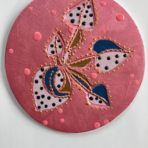 May include: A round, pink painted wooden ornament with a floral design. The flower has blue, gold, and black accents. The ornament has a small hole at the top for hanging. Pink dots are scattered around the floral design.