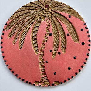 Könnte beinhalten: Ein rundes, korallenfarbenes Dekorationselement mit einem goldfarbenen Palmenmotiv. Die Palme befindet sich in der Mitte, mit detaillierten Wedeln und Stamm. Schwarze Punkte umranden den Rand. Oben befindet sich ein kleines Loch zum Aufhängen.
