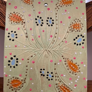 Può includere: Una piastrella quadrata in ceramica verde chiaro con un motivo floreale. Il design presenta forme floreali arancioni, nere e blu, insieme a punti rosa e bianchi. La piastrella ha un foro nella parte superiore per appenderla.