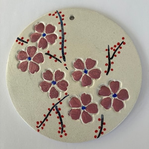May include: A round ceramic ornament with a white background and pink flowers with blue centers. The flowers are painted on the ornament and have black stems with red dots.