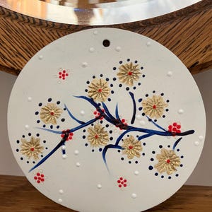 May include: A round, white ceramic ornament with a floral design. The design features gold-colored flowers with red centers, blue branches, and small red berries. The background has small white dots.