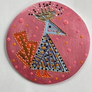 Puede incluir: Adorno redondo de madera pintada de color rosa con un diseño de pájaro estilizado. El pájaro tiene una cabeza azul, un cuerpo morado y azul y patas naranjas. El adorno está decorado con puntos dorados, negros y naranjas.