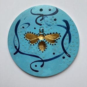 May include: A round, blue painted wooden ornament with a gold bee design and black swirls. The ornament is decorated with white dots.