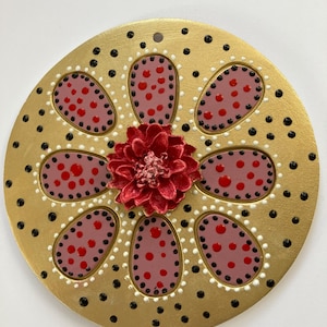 May include: A round, gold-painted wooden ornament with a red flower in the center. The flower is surrounded by six pink petals with red dots. The background is covered in black and white dots.