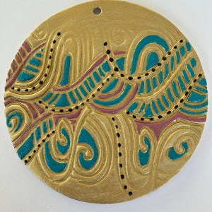 May include: A round, gold-colored ornament with a teal and pink abstract design. The ornament has a hole at the top for hanging.