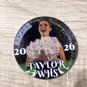 May include: A round button featuring a cheerleader with white pom-poms. The button has the text "SENIOR 2026 TAYLOR WHS" in white and blue. The background is a dark blue with white sparkles and a green field.