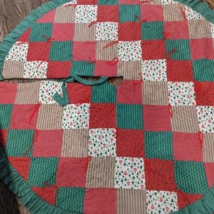 May include: A circular Christmas tree skirt featuring a patchwork design of red, green, and beige fabric squares. The pattern includes solid colors, thin stripes, and a small tree print. It is finished with a green ruffled edge and red ties.