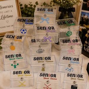 May include: A collection of clear acrylic keepsake boxes with hinged lids and metal latches. Each box features the text SENIOR 2026, a personalized name, and a unique graphic icon such as a sports ball, bow, or musical notes in various colors.