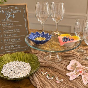 May include: A wine charm bar setup featuring a sign with instructions, four clear wine glasses, a glass pedestal tray with two small bowls containing beads, a green textured bowl filled with letter beads, and a bow-shaped dish with charms.
