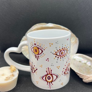 Puede incluir: Taza de cerámica blanca con asa en forma de corazón, decorada con un patrón repetido de ojos rojos, dorados y plateados. Los ojos tienen un diseño estilizado con un punto en el centro.