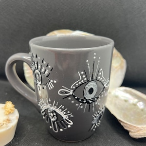 Puede incluir: Una taza de cerámica gris con un diseño de ojo blanco y negro. Los ojos son estilizados y tienen un patrón de puntos alrededor.