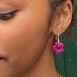Peut inclure: Gros plan sur une oreille ornée d'une boucle d'oreille dorée avec un pendentif fleur rose vif. La fleur a un centre circulaire et cinq pétales arrondis. La boucle d'oreille est au centre de l'image, sur un fond flou.