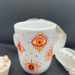 Puede incluir: Taza de cerámica blanca con un patrón de ojos naranja, amarillo y morado. Los ojos tienen un fondo blanco y están rodeados por un patrón radiante de puntos.