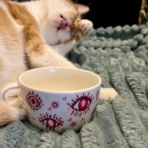 Puede incluir: Taza de cerámica blanca con un diseño de ojo rosa y rojo. La taza está sobre una manta gris peluda.
