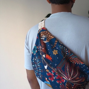 May include: A crossbody bag featuring a dark blue fabric with a vibrant jungle-themed print, including a leopard, a toucan, and various tropical flowers and leaves. The bag has a metallic zipper closure and a patterned woven strap across the shoulder.