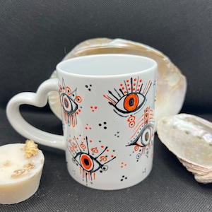 Puede incluir: Taza de cerámica blanca con asa en forma de corazón. La taza está decorada con un diseño de ojo negro y naranja.