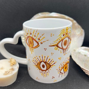 Puede incluir: Taza de cerámica blanca con asa en forma de corazón. La taza está decorada con ojos pintados en dorado y marrón con un diseño de rayos de sol.
