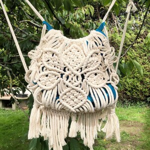 Könnte beinhalten: Eine weiße Makramee-Tasche mit Fransen, die an einem Baumzweig hängt. Die Tasche hat ein blaues Futter.