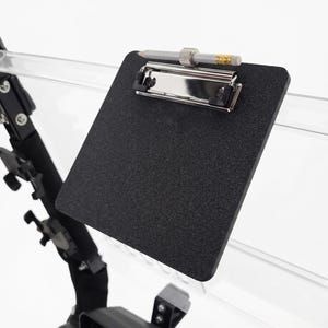 May include: A black square clipboard with a textured surface and a silver metal clip mechanism. A grey pencil with a silver ferrule and pink eraser is resting horizontally across the top of the clip. The clipboard is attached to a clear stand.