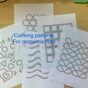 Può includere: Diversi fogli di carta bianca con disegni a tratto nero di forme e motivi geometrici. Il testo "Calming patterns For dementia care" è stampato sui fogli. Sullo sfondo sono visibili pennarelli colorati.