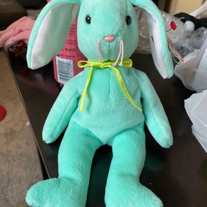 Puede incluir: Un conejito de peluche verde claro con orejas blancas y un lazo amarillo. El conejito tiene la nariz rosa y los ojos negros.