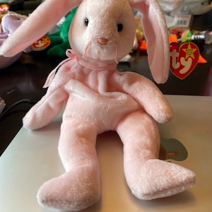 May include: Pink plush bunny rabbit toy with a pink ribbon around its neck. The toy has a Ty tag attached to its ear with the words "Beanie Baby" on it.