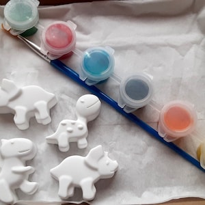 May include: A set of six paint pots with a blue paintbrush, and four white ceramic dinosaur figurines. The paint colors are green, red, blue, gray, orange, and yellow.