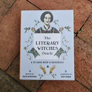 以下が含まれることがあります： 「The Literary Witches Oracle」という名前の70枚のオラクルカードデッキが入った青い箱で、白い縁取りがされています。箱には、黒いドレスを着た黒髪の女性が花と鳥に囲まれているイラストが描かれています。箱には「Written by Taisia Kitaiskaia」と「Illustrated by Katy Horan」の文字が印刷されています。