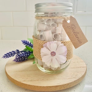 Puede incluir: Un tarro de vidrio lleno de papeles enrollados, decorado con una cinta de arpillera, una flor blanca con un centro rosa y una etiqueta marrón que dice "Jilanie".