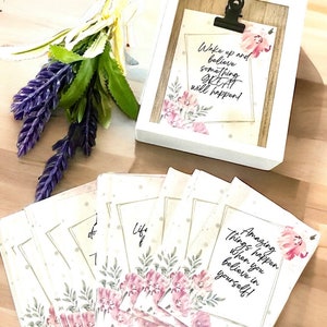 May include: A set of eight white cards with floral designs and inspirational quotes. The quotes include "Life is..." and "Amazing things happen when you believe in yourself!" The cards are arranged in a fan shape on a wooden surface.