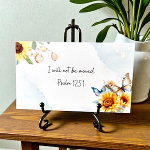 May include: A decorative card with the text "I will not be moved. Psalm 125:1" displayed on a black metal easel. The card features watercolor sunflowers and butterflies with a light blue background.