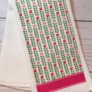 May include: White kitchen towel with a pink and green floral print, a pink ribbon, and white lace trim.
