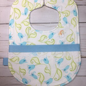 May include: A white baby bib with a blue floral pattern, a blue ribbon, and white lace trim. The bib has two white snaps on the neck.