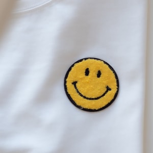 Könnte beinhalten: Ein weißes T-Shirt mit einem gelben Smiley-Aufnäher. Der Smiley hat einen schwarzen Umriss und schwarze Augen und Mund. Das Shirt besteht aus einem weichen, glatten Stoff.