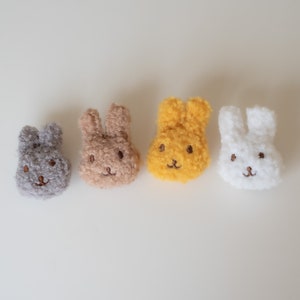 Könnte beinhalten: Vier Plüsch-Hasengesichter in einer Reihe, jeweils mit gestickten Details. Die Hasen sind in Grautönen, Beige, Gelb und Weiß gehalten. Die Gesichter haben kleine, gestickte Augen und ein aufgenähtes Lächeln.