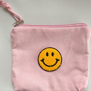 Könnte beinhalten: Eine rosa Canvas-Kosmetiktasche mit einem aufgestickten gelben Smiley auf der Vorderseite.
