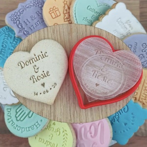 May include: A set of heart-shaped biscuits with personalised messages, including "Dominic & Rosie" and "Dominic & Rosie" with a date. The biscuits are surrounded by other decorated biscuits with various designs and messages.