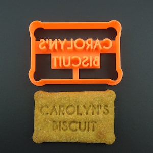 May include: Orange plastic cookie cutter with the text "CAROLYN'S BISCUIT" in reverse. A biscuit with the same text imprinted on it is shown below the cutter.