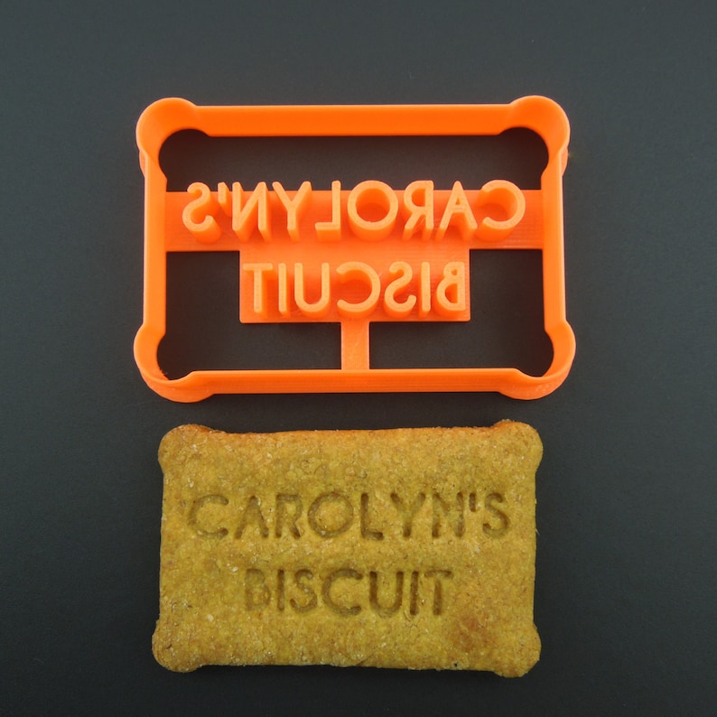 May include: Orange plastic cookie cutter with the text "CAROLYN'S BISCUIT" in reverse. A biscuit with the same text imprinted on it is shown below the cutter.