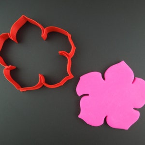 Peut inclure: Un emporte-pièce en plastique rouge en forme de fleur, à côté d'un morceau de fondant rose en forme de fleur.