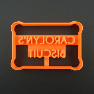May include: Orange plastic cookie cutter shaped like a rectangle with the words "CAROLYN'S BISCUIT" embossed on it.