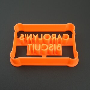 May include: Orange plastic cookie cutter with the words "CAROLYN'S BISCUIT" cut out.