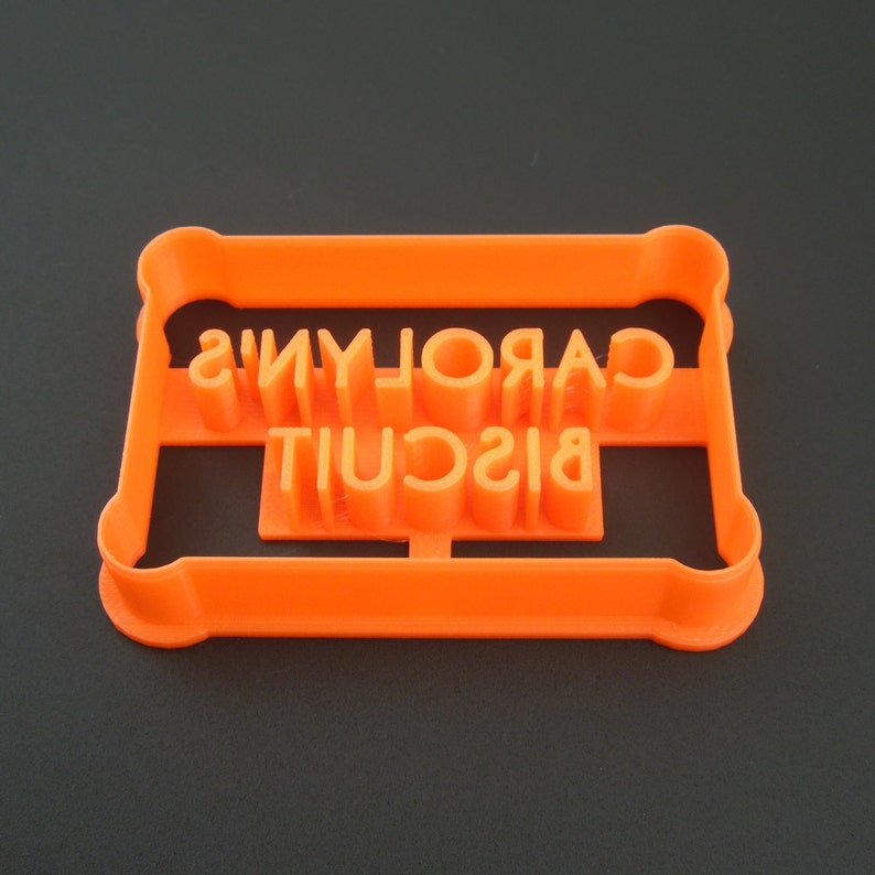 May include: Orange plastic cookie cutter with the words "CAROLYN'S BISCUIT" cut out.