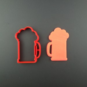 May include: Two red plastic cookie cutters shaped like beer steins. One cutter is solid and the other has an outline of the stein.
