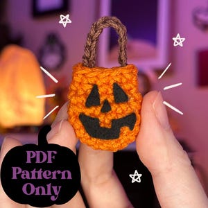 May include: An orange crocheted pumpkin-shaped bag with a black face and a brown handle. The bag is held in a person's hand. The text "PDF Pattern Only" is in a black pumpkin shape.