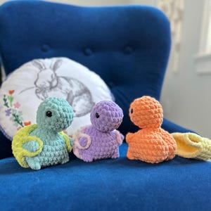 May include: Three small, plush, crocheted turtle toys in light green, lavender, and orange. Each turtle features a rounded shell, a textured yarn body, and small black bead eyes. A separate yellow crocheted shell piece sits to the right.