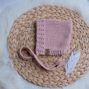 May include: A light pink knitted baby bonnet with a braided tie and tassel. The bonnet is placed on a woven placemat with a white feather with gold accents. The bonnet has a small leather tag with the initials "WF".