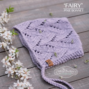May include: A lilac knitted pixie bonnet with a textured pattern. The bonnet features a braided tie and a small leather tag with the initials "WF". The text "FAIRY" and "PIXIE BONNET" are displayed above the bonnet. White Feather Handmade is also displayed.