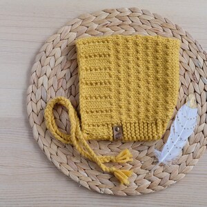 May include: A yellow knitted baby bonnet with a long tie. The bonnet has a small brown leather tag with the text "Wee WF" on it.